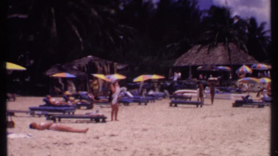 1969 Bahamas beach resort view with sunbathers, lounge chairs, colorful umbrellas, thatched cabanas, and palms in soft 8mm color.