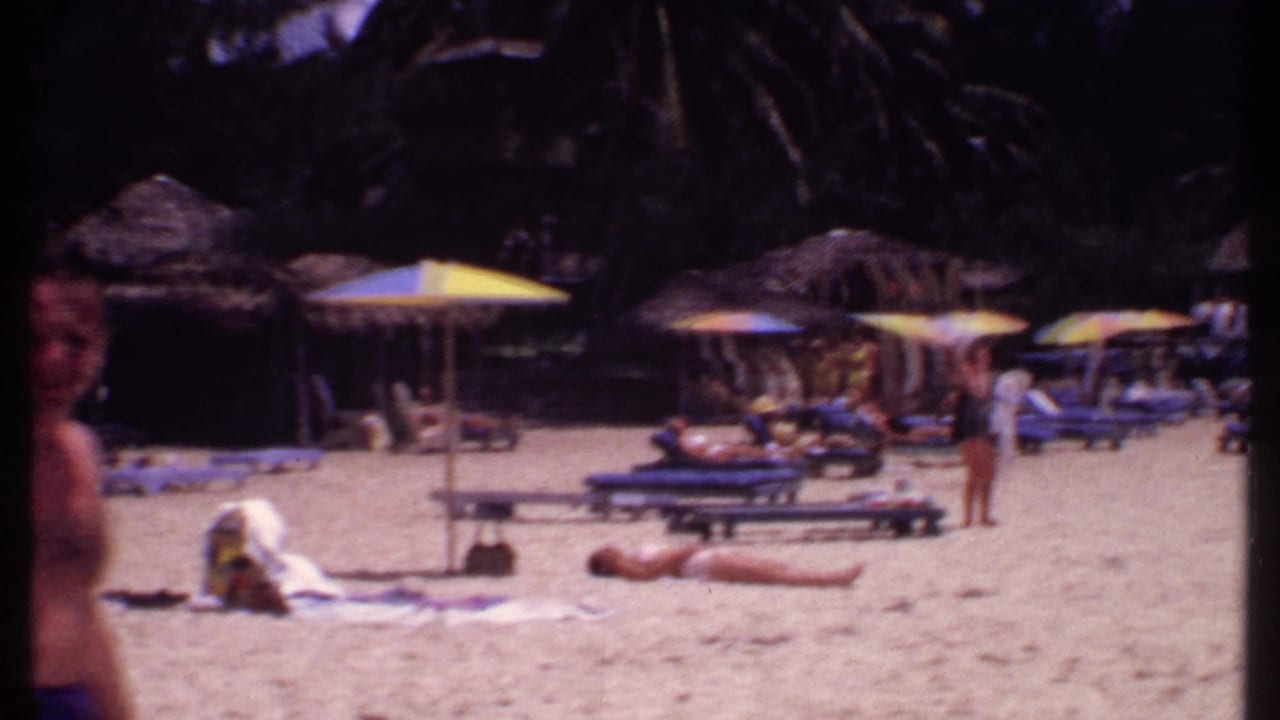 Sunbathers and lounge chairs line a sandy beach with umbrellas and palm trees behind them.