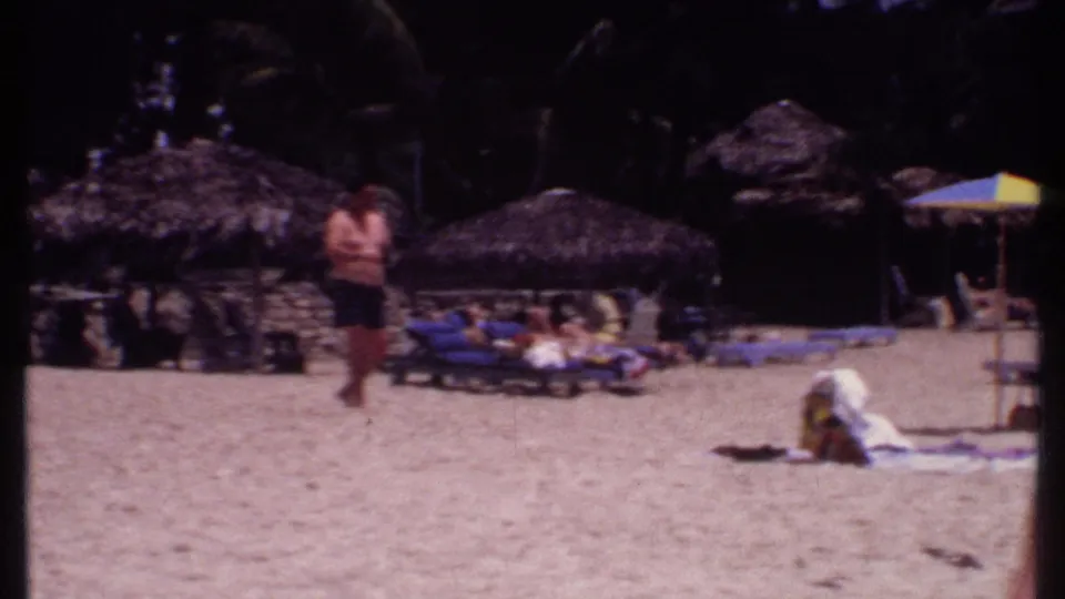 1969 Bahamas beach resort frame showing a visitor walking past loungers, towels, umbrellas, thatched cabanas, and dark palms.
