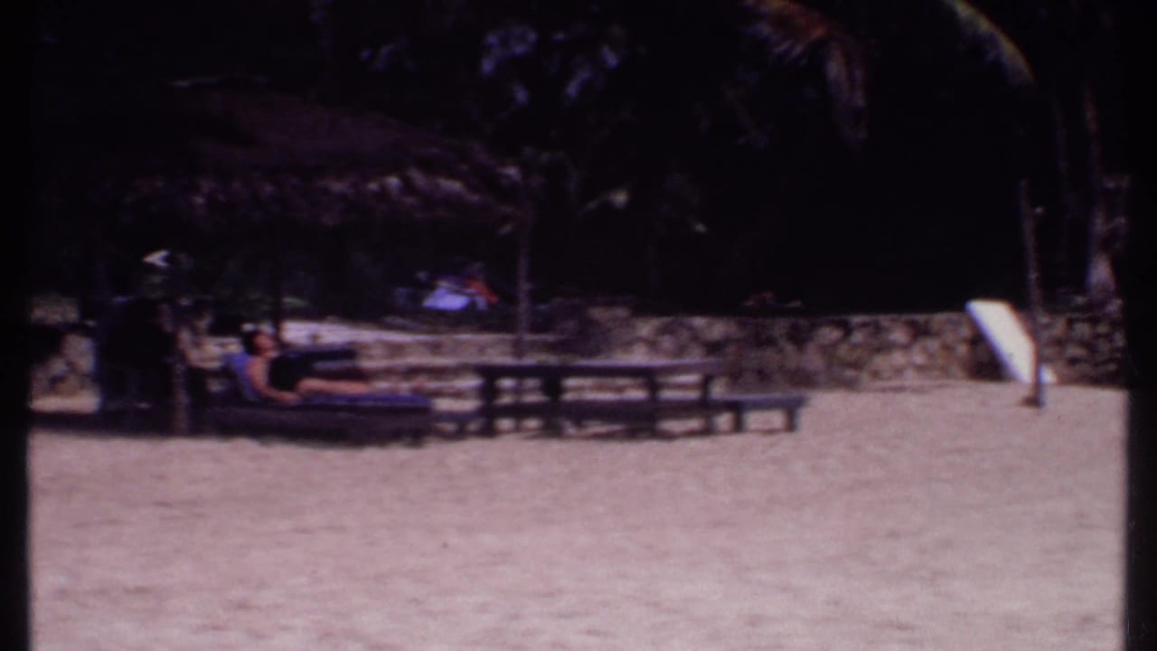 1969 Bahamas resort view with sandy ground, palm shade, picnic tables, and distant visitors under a thatched structure.