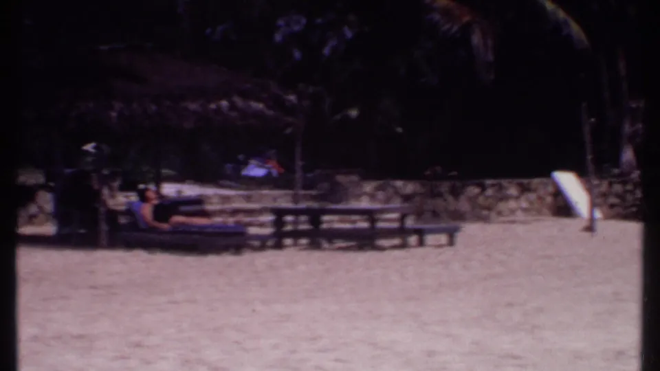 1969 Bahamas resort view with sandy ground, palm shade, picnic tables, and distant visitors under a thatched structure.