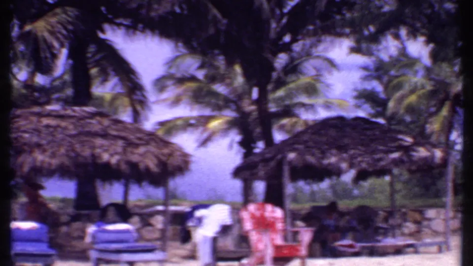 1969 Bahamas resort loungers beneath thatched cabanas, with palms, a low wall, and water visible beyond the shade.