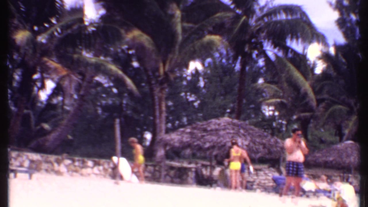 1969 Bahamas 8mm frame showing beachgoers near thatched cabanas, palm trees, and pale sand.