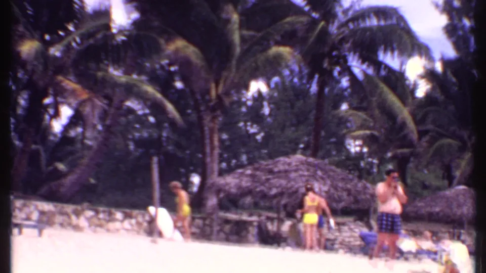 1969 Bahamas 8mm frame showing beachgoers near thatched cabanas, palm trees, and pale sand.