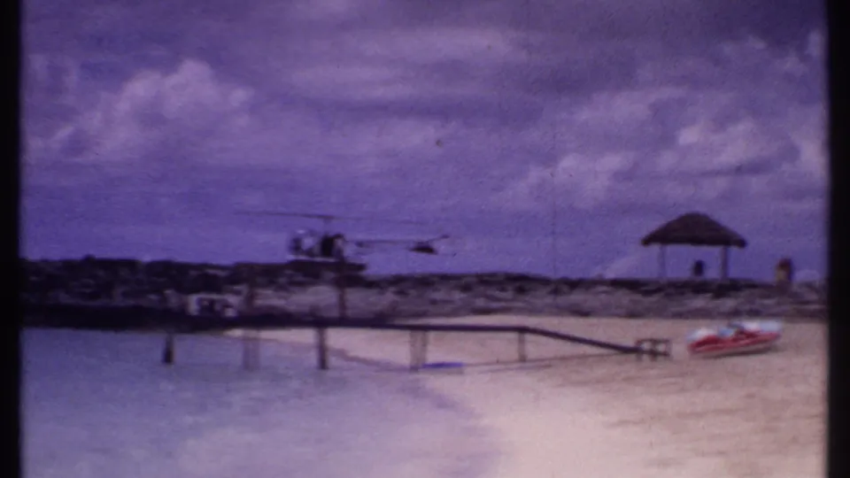 1969 Bahamas beach view with shallow water, a small pier, thatched shelter, distant helicopter, and boat on sand.