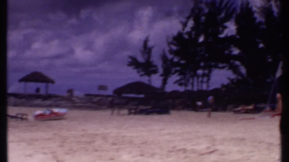 1969 Bahamas 8mm beach frame with thatched shelters, palm trees, a red-and-white boat, and scattered visitors.