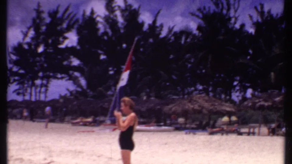 1969 Bahamas beach frame showing a woman in black swimwear standing near thatched cabanas and palm trees.