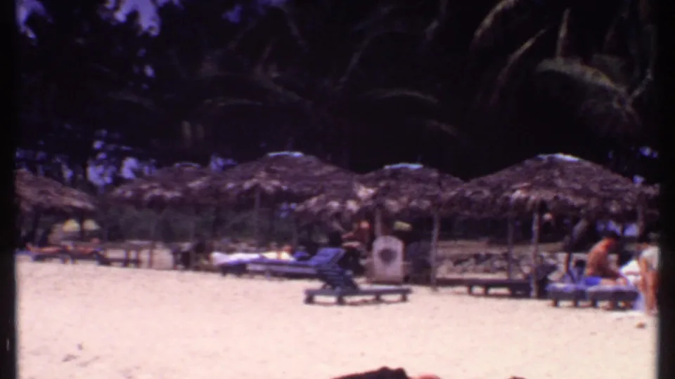 1969 Bahamas beach view with thatched shade huts, palm trees, lounge chairs, and vacationers scattered across the sand.