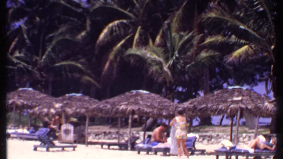 1969 Bahamas resort beach frame showing palm trees, thatched shade huts, lounge chairs, and people standing or reclining in swimwear.