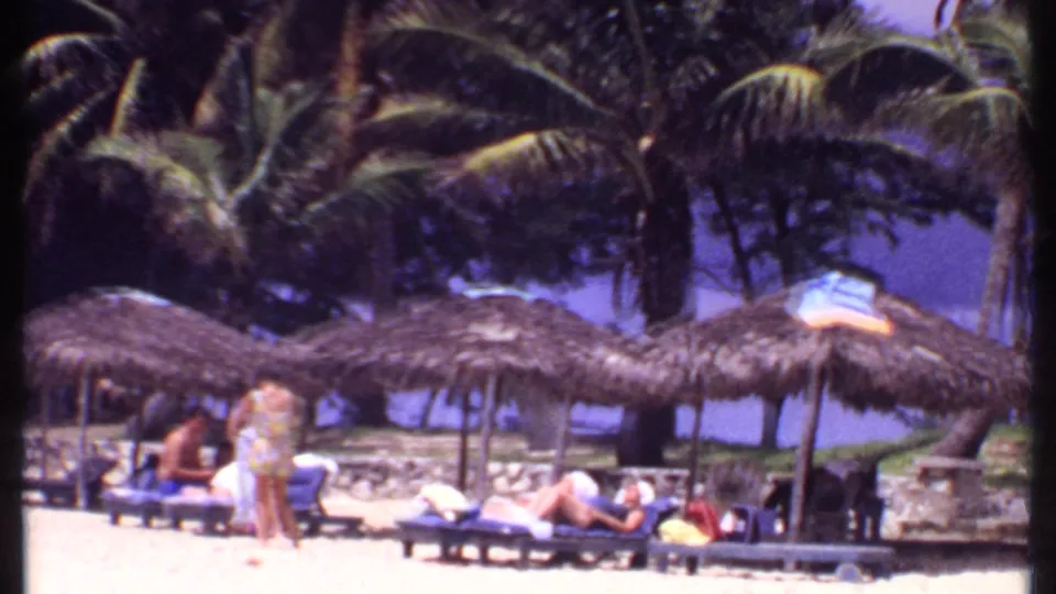 1969 Bahamas beach frame with vacationers on lounge chairs under thatched shade huts, backed by palm trees and bright sand.