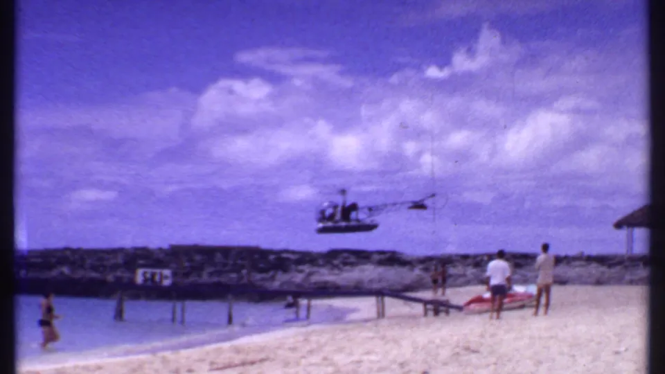 1969 Bahamas beach scene with a helicopter passing low over swimmers, people on the sand, a rocky shoreline, and a visible shore sign.