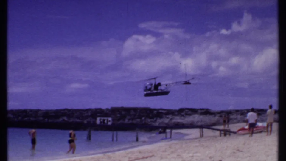 1969 Bahamas shoreline view showing a low helicopter, swimmers near the beach, standing beachgoers, clouds, and a visible sign by the water.
