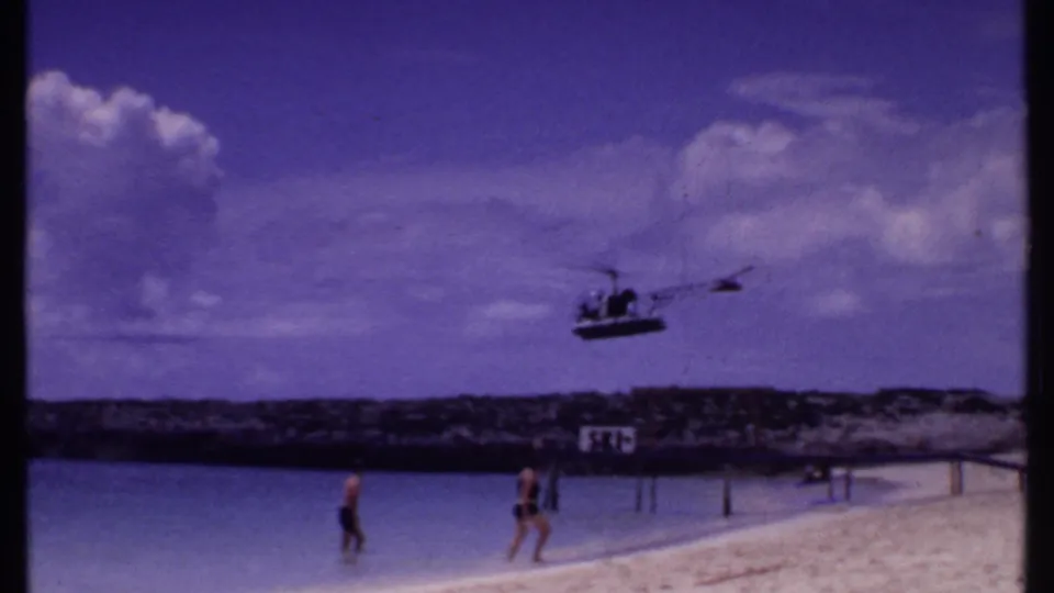 1969 Bahamas beach footage with a helicopter overhead, two people near the waterline, a cloudy sky, and a rocky shore behind them.