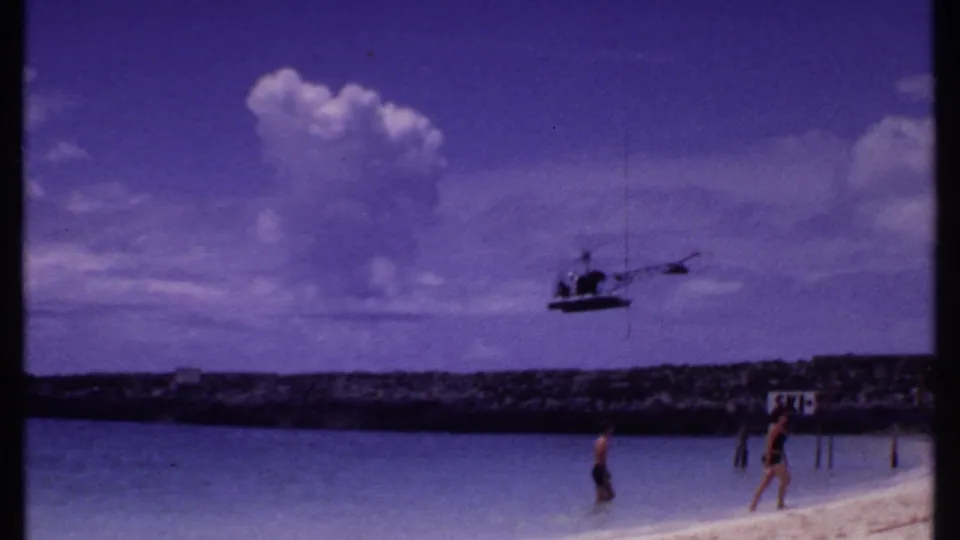 1969 Bahamas beach frame showing a helicopter above the shore, two beachgoers in shallow water, clouds, and a visible sign near the rocky bank.