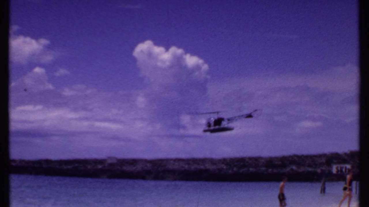 Helicopter flying over blue ocean near a rocky shoreline with beachgoers at the lower edge.