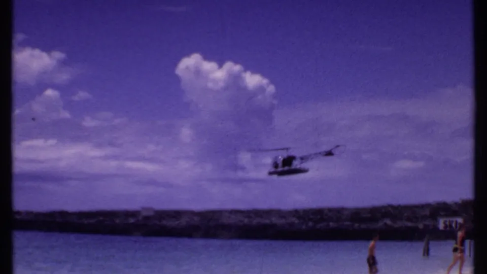 1969 Bahamas 8mm frame showing a helicopter over turquoise water, distant rocky barrier, clouds, and a few beachgoers near the shore.