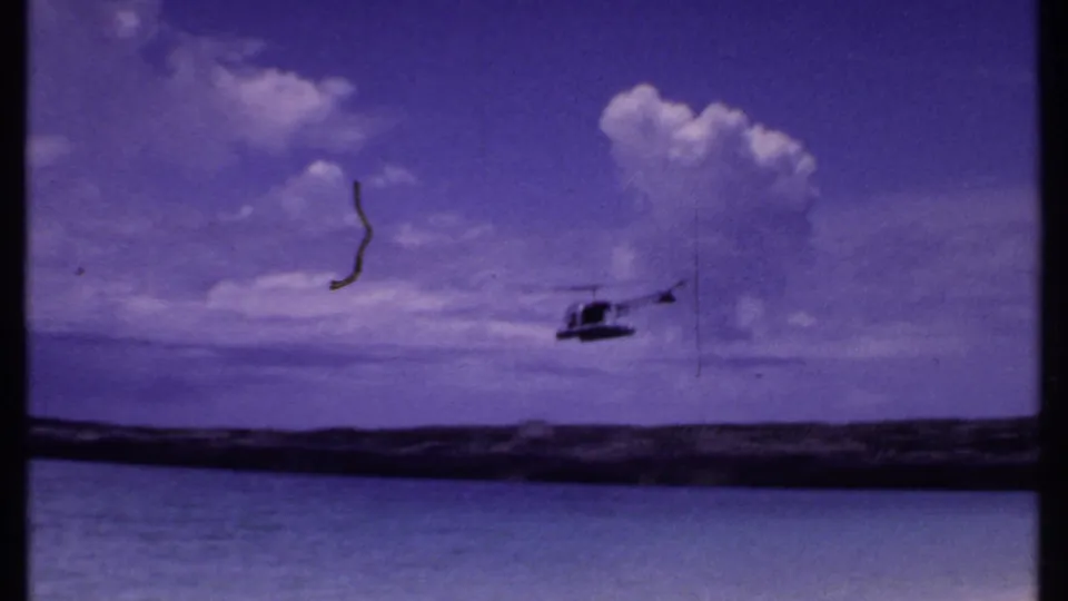 1969 Bahamas 8mm frame with a helicopter flying above ocean water and a rocky barrier beneath a purple-tinted cloudy sky.
