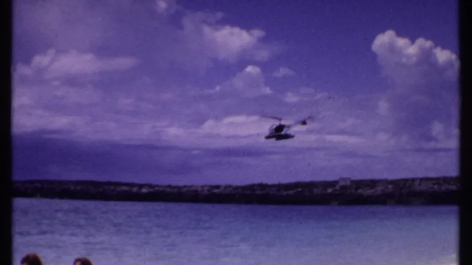 1969 Bahamas 8mm frame showing a small helicopter over the sea, with a low rocky shore and broad cloud cover behind it.