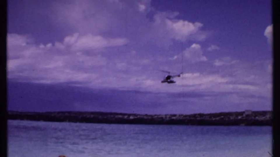 1969 Bahamas 8mm frame of a helicopter near the horizon, crossing above calm ocean water and a low rocky shoreline.