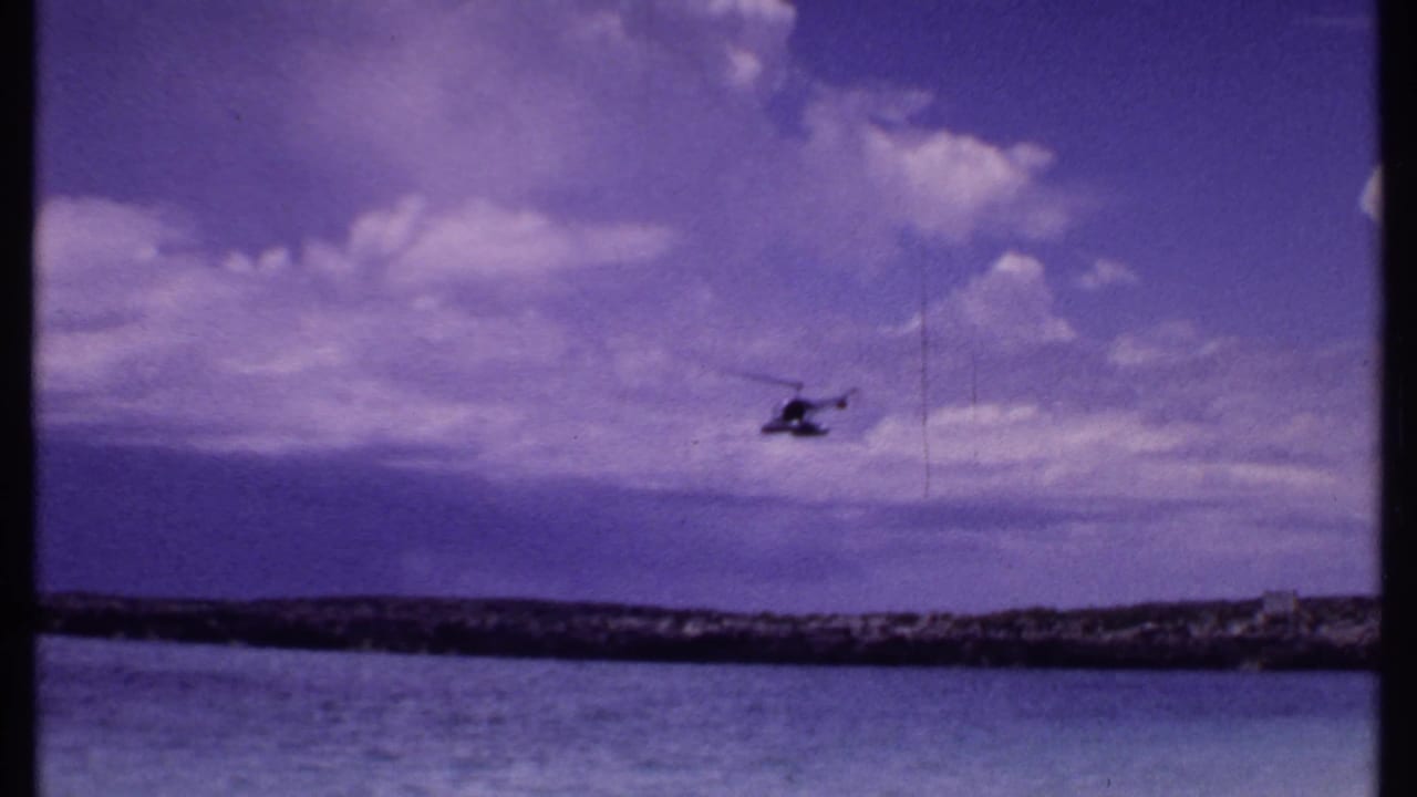 1969 Bahamas 8mm frame showing a distant helicopter over calm water, low shoreline, and large white clouds in a blue-purple sky.