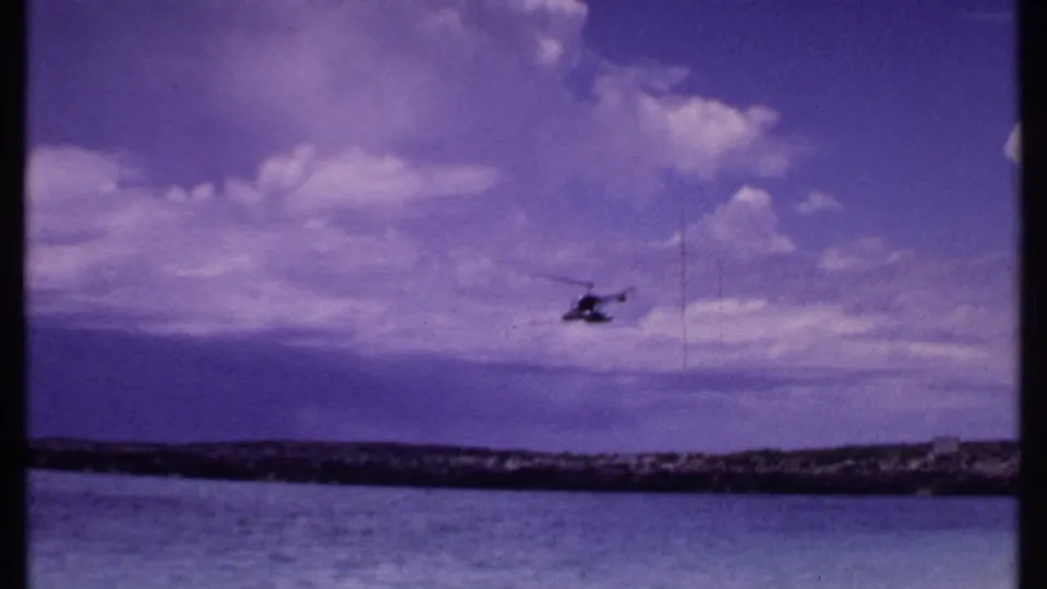 1969 Bahamas 8mm frame showing a distant helicopter over calm water, low shoreline, and large white clouds in a blue-purple sky.