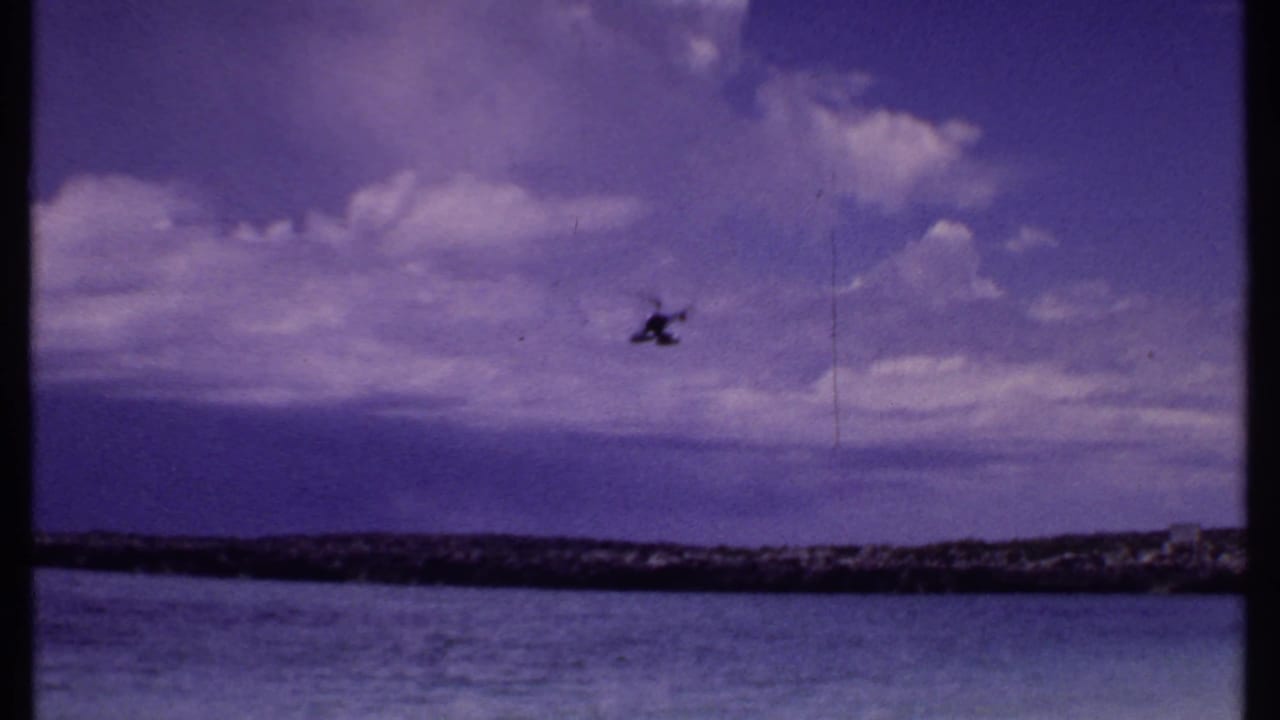 Distant helicopter in open sky above a low shoreline and water, with clouds behind it.
