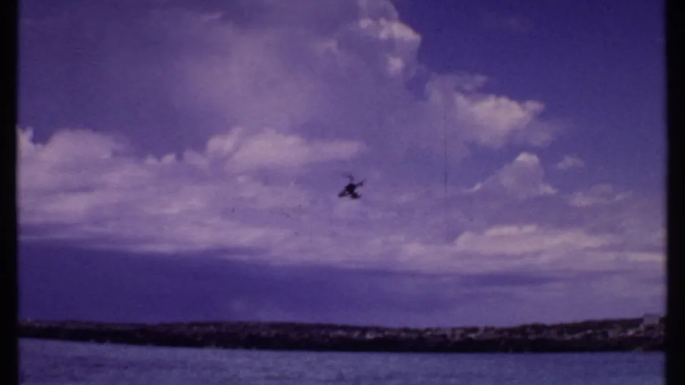 1969 Bahamas 8mm frame showing the helicopter as a small silhouette against bright clouds over coastal water.