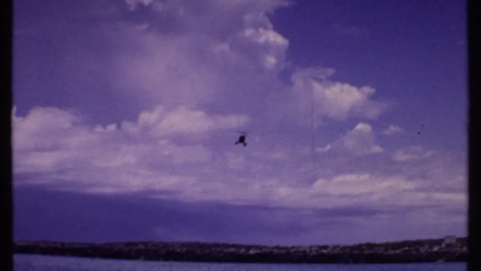 1969 Bahamas home-movie frame with a tiny helicopter in a broad sky, above a dark coastal strip and calm water.
