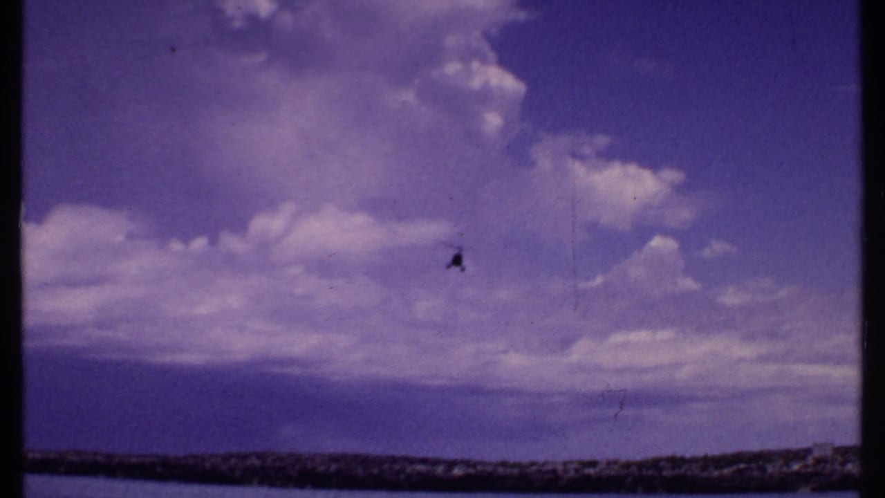 1969 Bahamas home-movie frame with a small helicopter flying over clouds and a low coastal horizon.
