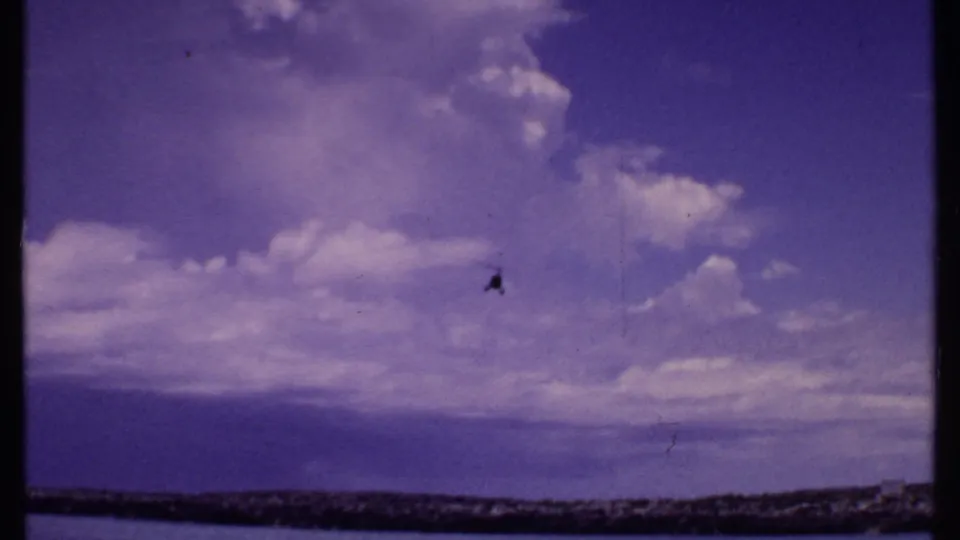 1969 Bahamas home-movie frame with a small helicopter flying over clouds and a low coastal horizon.