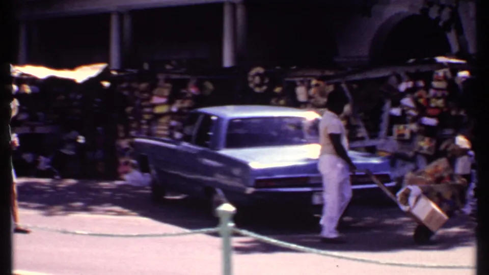 1969 Bahamas street-market scene with pedestrians near stalls, a light blue classic car, and a man pushing a cart.