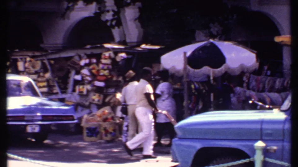 1969 Bahamas street scene with people near market displays, umbrellas, carts, and blue classic cars.