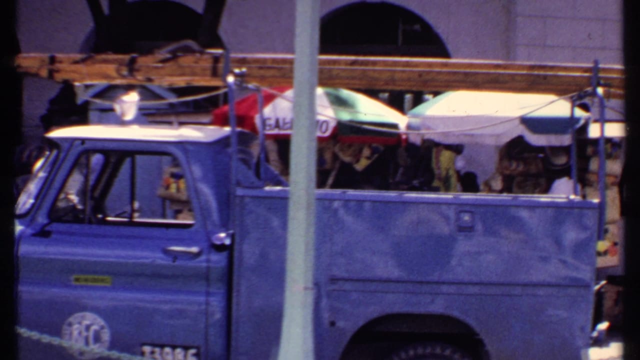 Side view of a blue truck passing market stalls shaded by umbrellas.