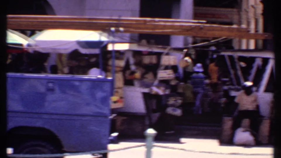 The rear of the blue truck frames a Bahamas market area with shaded stalls, pedestrians, hanging goods, and bright pavement.