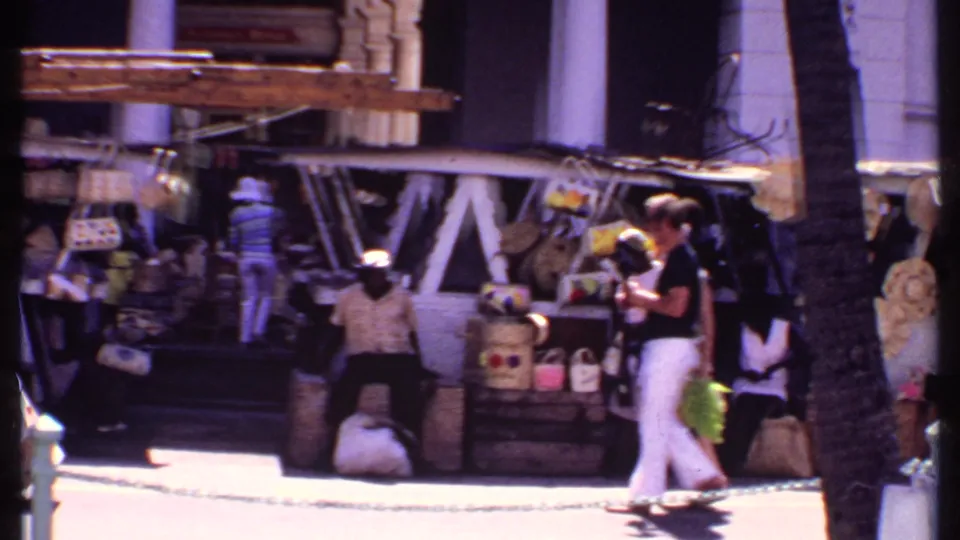 At a Bahamas street market, pedestrians pass vendor displays with bags, baskets, hanging items, a palm trunk, and shaded storefronts.