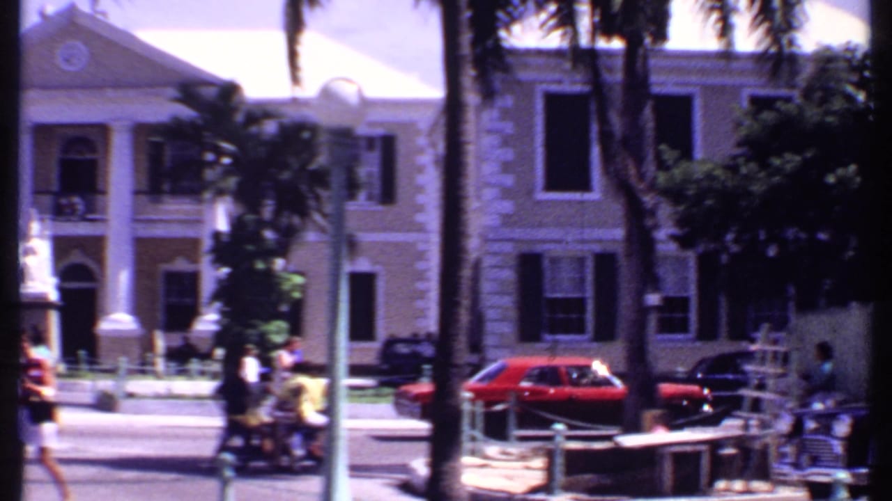 1969 Bahamas street view with pedestrians, palm trees, a red car, and a colonial-style building with white columns.