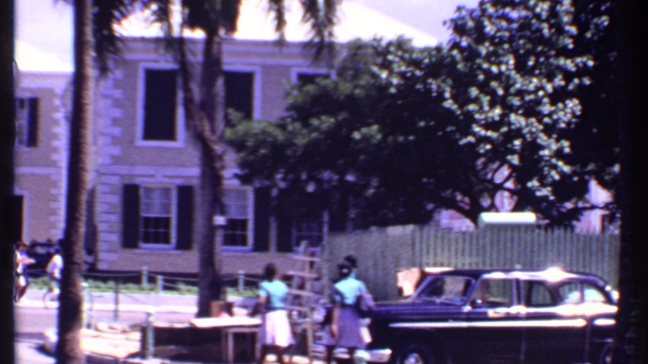 Dark sedan and pedestrians along a sunlit street with palms and a shuttered building behind them.