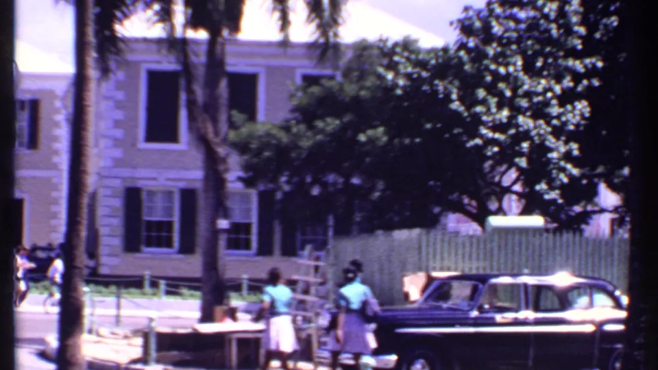 1969 Bahamas street scene with a dark sedan, pedestrians, palm trunks, fencing, and a shuttered building in bright sun.