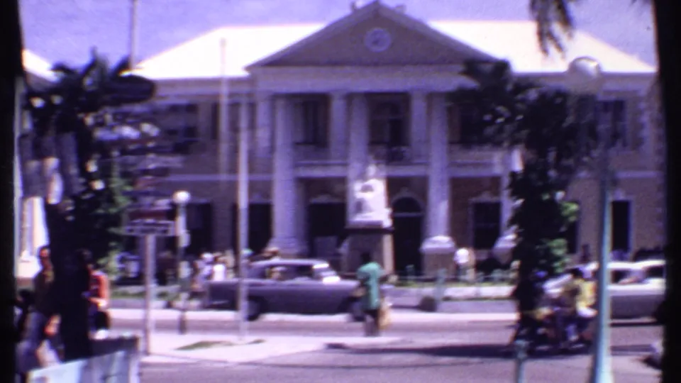 1969 Bahamas view across a street toward a columned public-looking building, pedestrians, a flagpole, and classic cars.