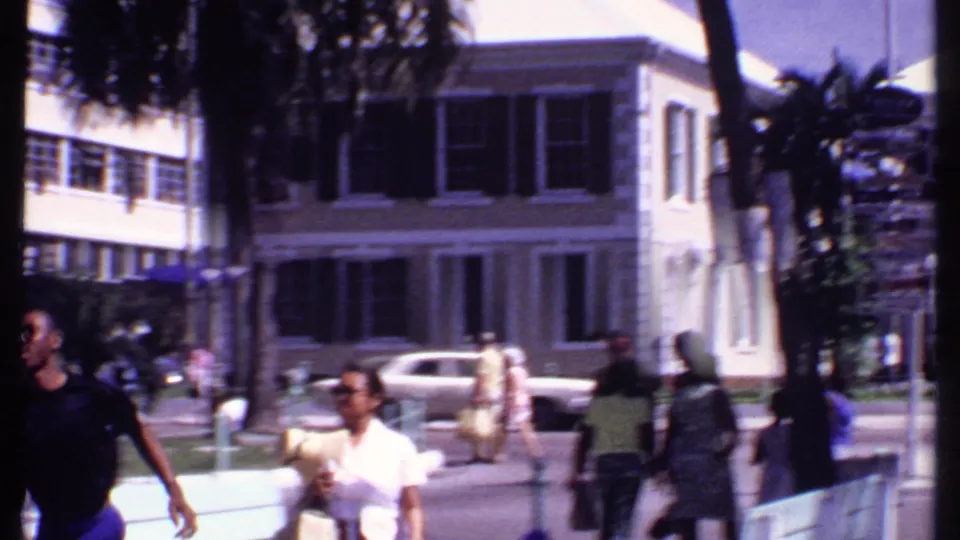 1969 Bahamas street view with pedestrians in the foreground, classic cars, palm trees, and a shuttered building across the road.