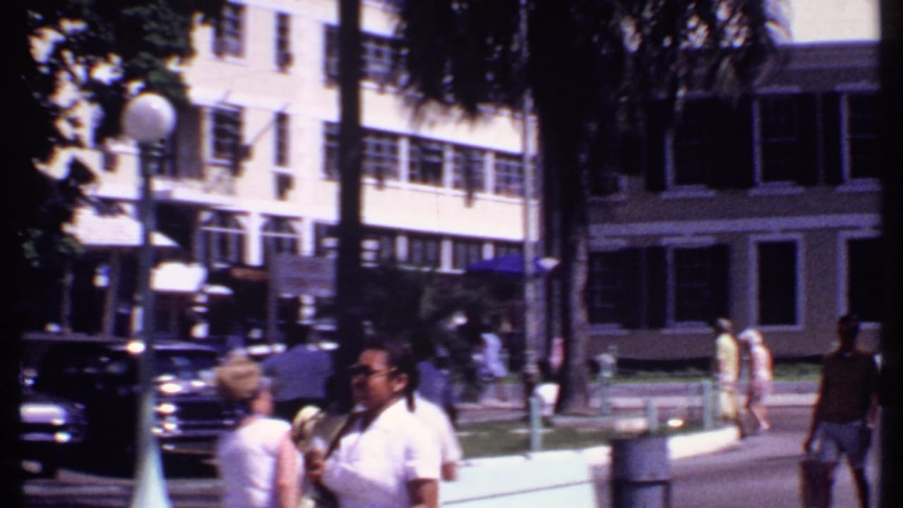Pedestrians walk near parked cars, palm trees, and shuttered buildings on a sunny downtown street.