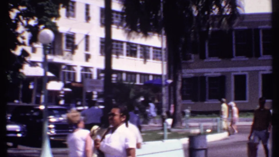 1969 Bahamas downtown street view with pedestrians in light clothing, classic cars, palm trees, and shuttered building facades.