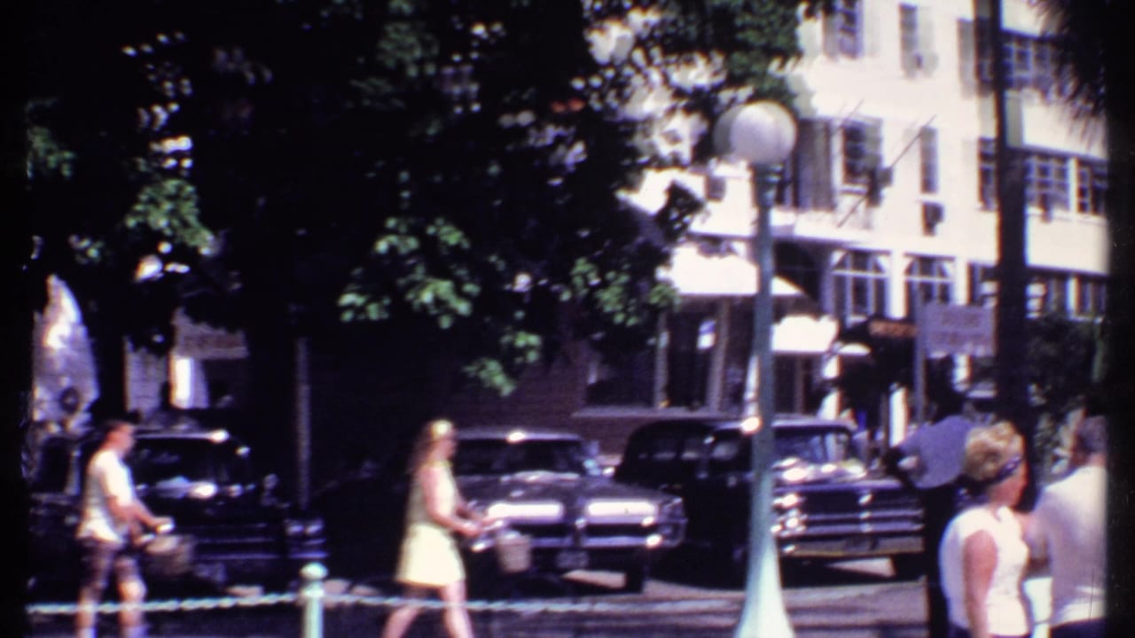 Pedestrians pass parked cars and shaded storefronts beneath trees on a downtown street.