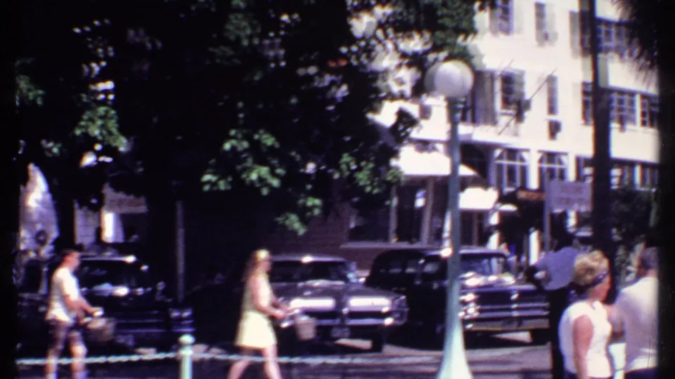 1969 Bahamas street scene with walkers, classic cars, tree shade, storefront awnings, and multi-story shuttered buildings.