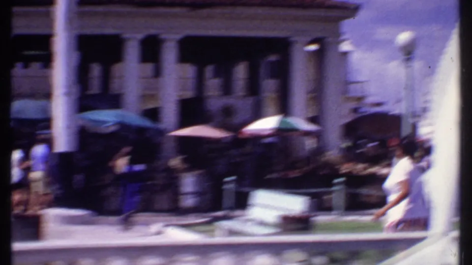 Soft 8mm frame of a Bahamas commercial street, with columns, awnings, umbrellas, pedestrians, and motion blur from handheld filming.
