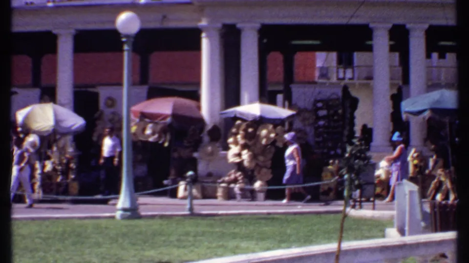 1969 Bahamas downtown commerce scene with market umbrellas, outdoor goods, pedestrians, columns, a low fence, and a globe street lamp.
