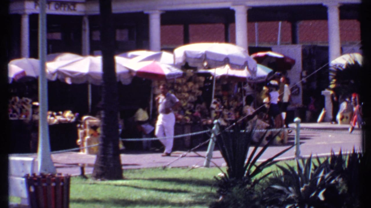 Man in light shirt and white trousers near vendor umbrellas and market goods.