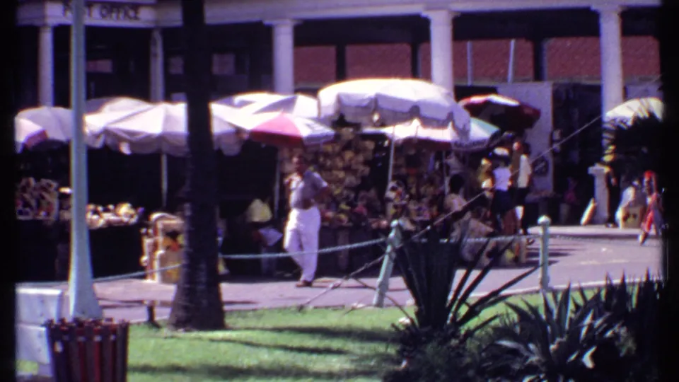1969 Bahamas market frame showing a man walking near shaded vendor displays, storefront columns, palms, and passersby.