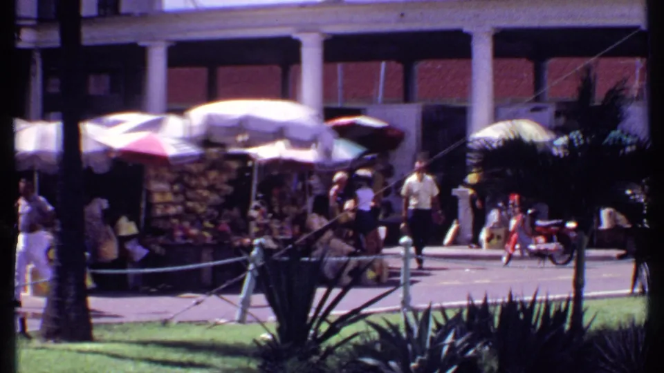 1969 Bahamas street market scene with umbrella stalls, people walking and sitting, palm trunks, plants, and shaded storefronts.