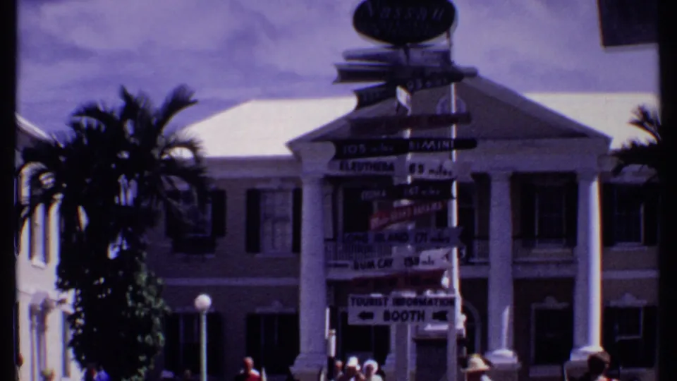 1969 Bahamas home-movie frame showing a tourist signpost, palm trees, and a columned building in a plaza.